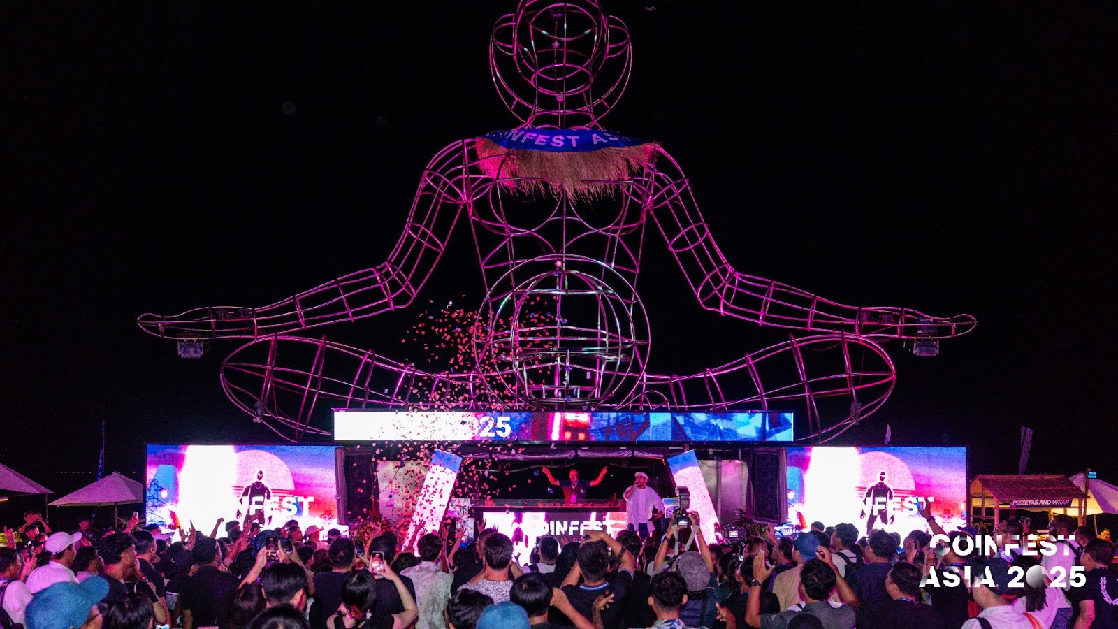 Nighttime concert and show at Coinfest Asia 2025 in Bali, featuring a glowing human-shaped statue and a lively crowd.